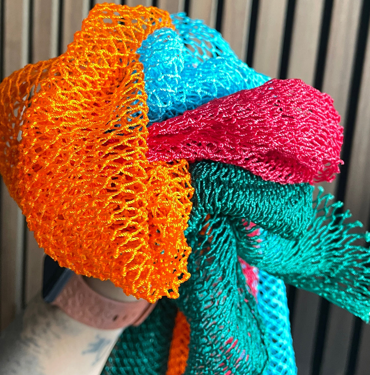 A close-up of a multicolor open-knit mesh fabric accessory African Net sponge, showing bright orange, turquoise blue, pink, and teal sections draped over a person's arm against a vertical wood slat background.