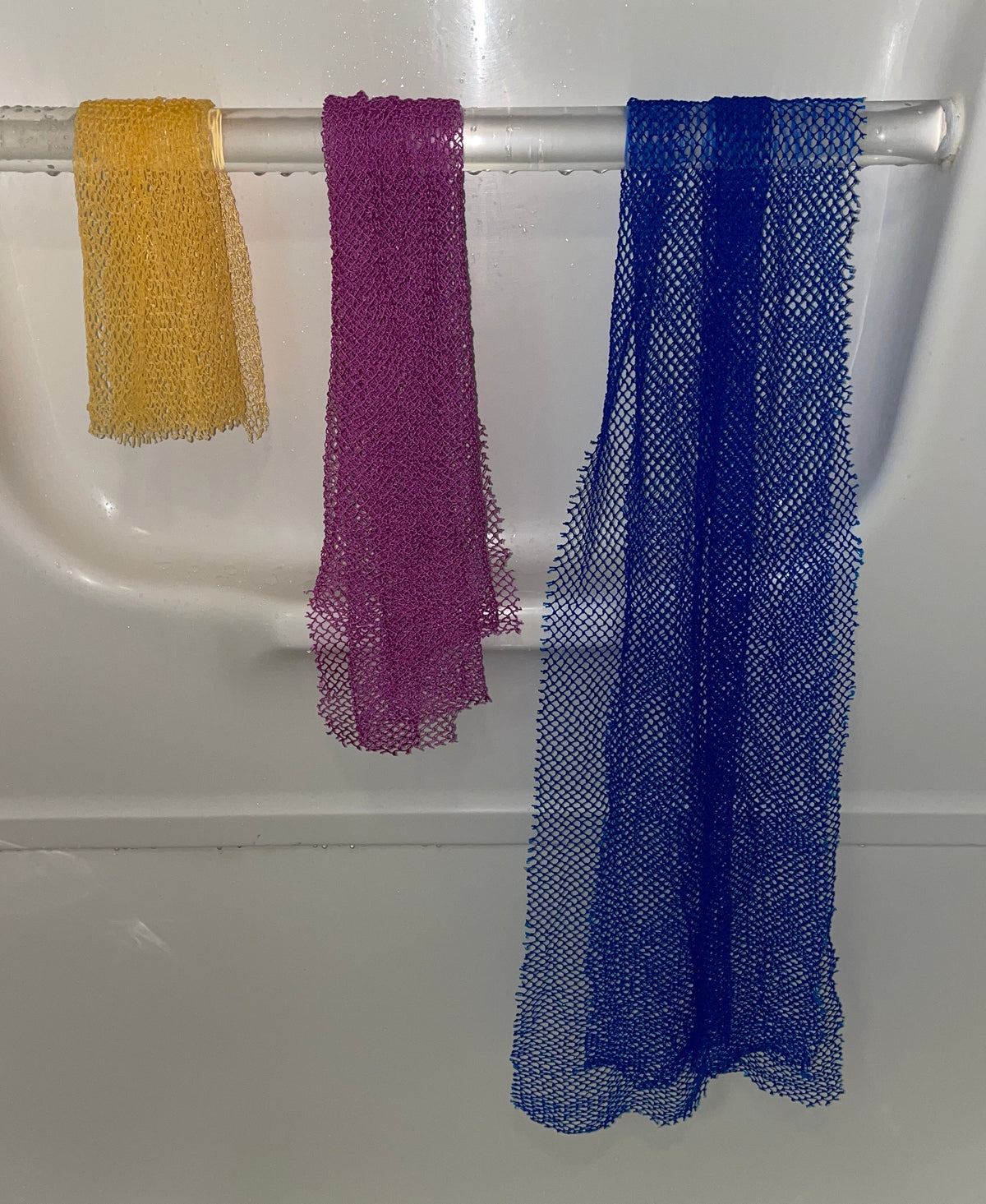 Three African Net sponges (mesh washcloths) in yellow, purple, and blue hanging from a white tub rod, showcasing 3 different sizes.
